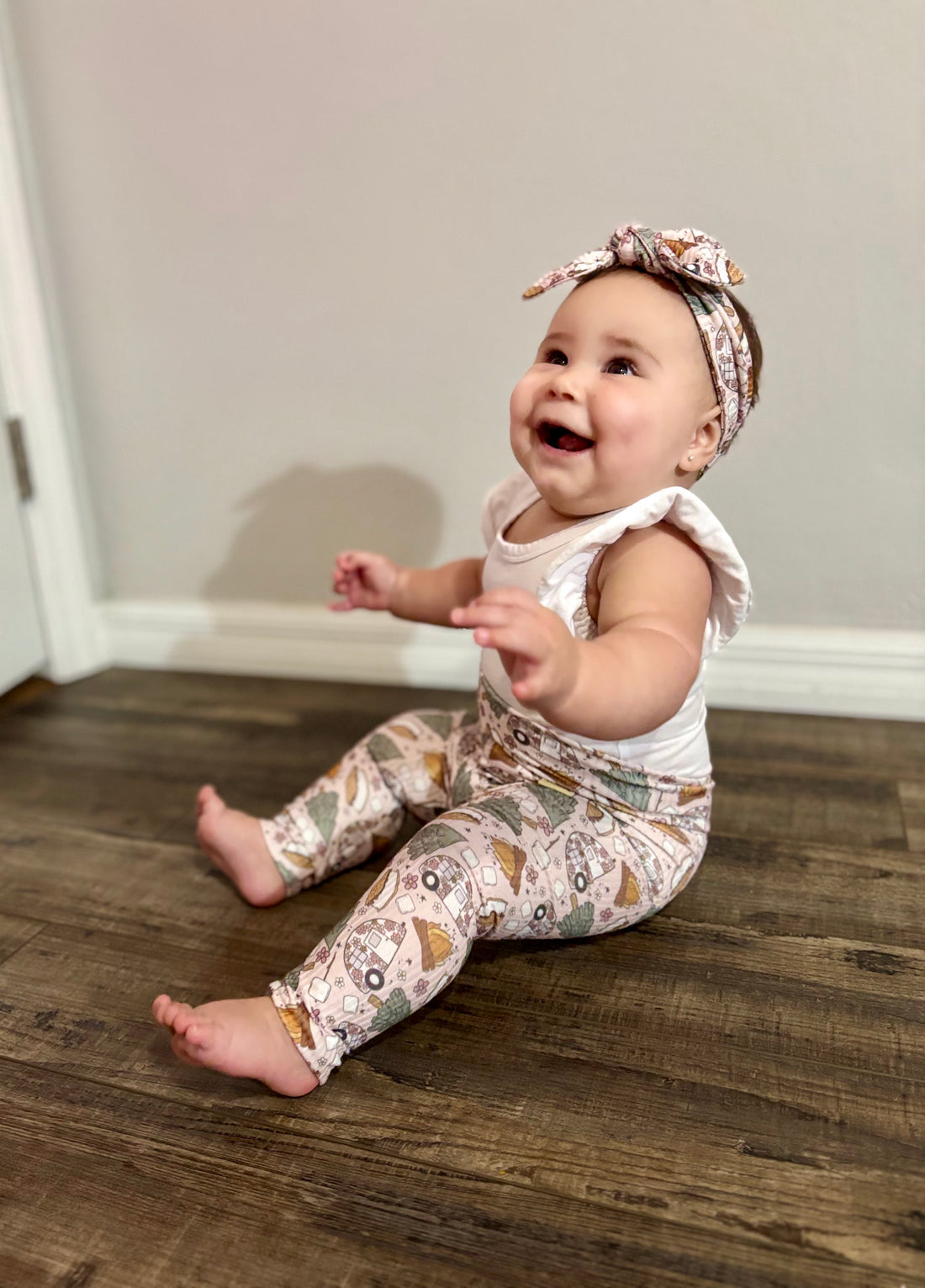 Baby sitting on a wooden floor wearing a white top and  matching camping print leggings and headband.