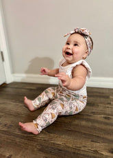 Baby sitting on a wooden floor wearing a white top and  matching camping print leggings and headband.