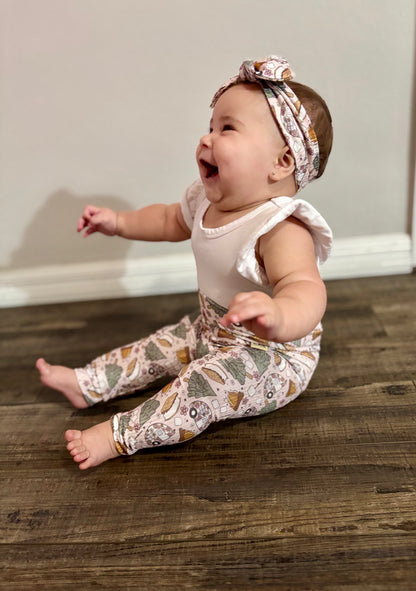 Baby sitting on a wooden floor wearing a white top and  matching camping print leggings and headband.