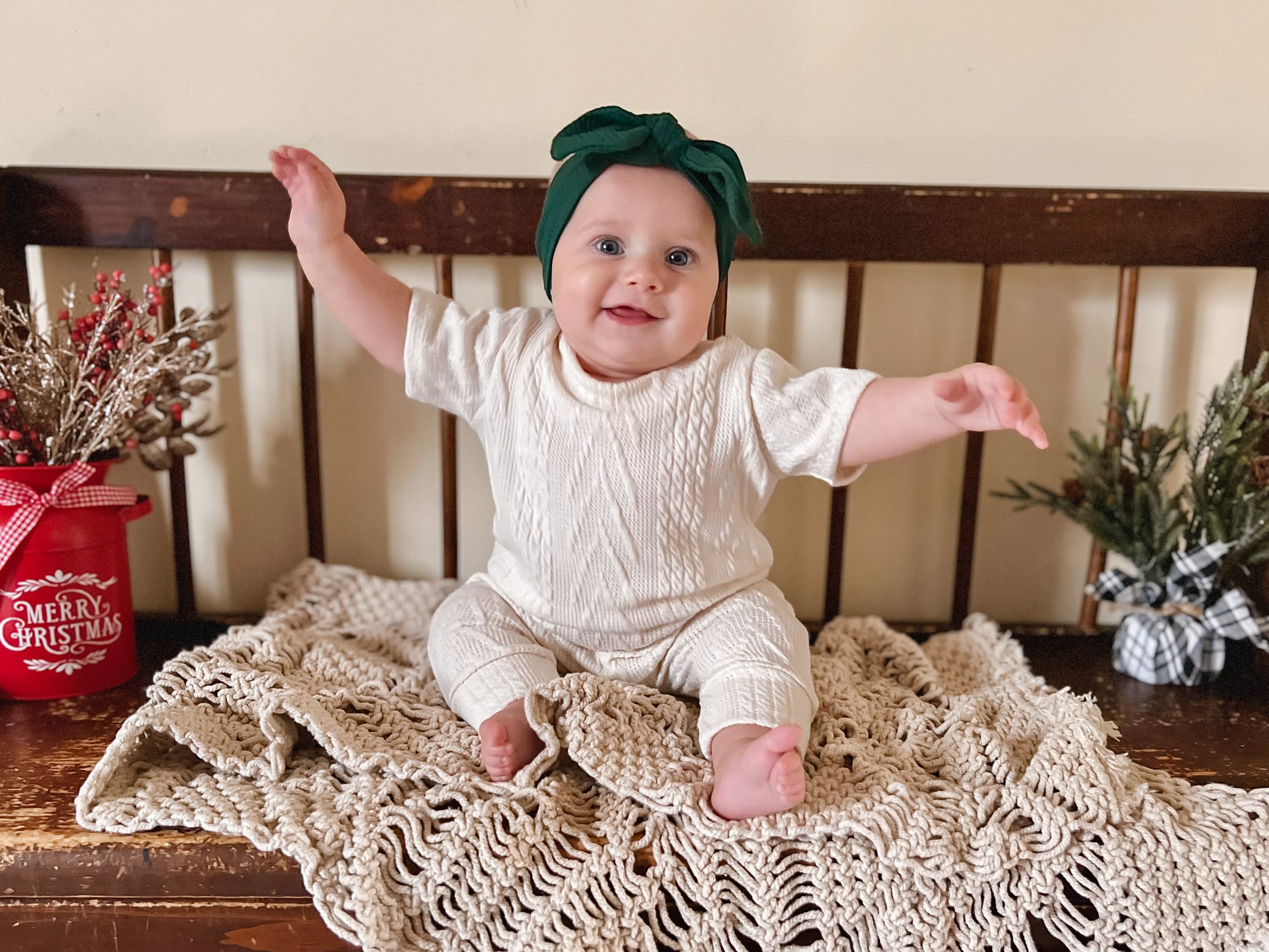 Baby sitting on a wooden bench with a knitted blanket, wearing a green headband and cream knit romper.