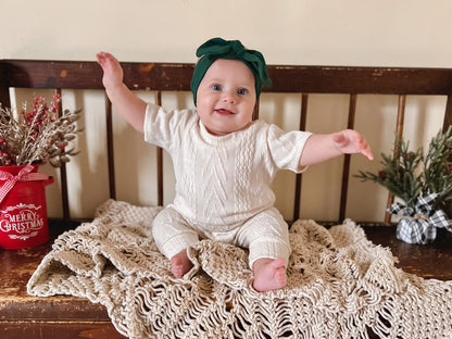 Baby sitting on a wooden bench with a knitted blanket, wearing a green headband and cream knit romper.