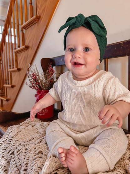 Baby wearing a green headband and beige harem romper sitting on a textured surface with a staircase in the background.