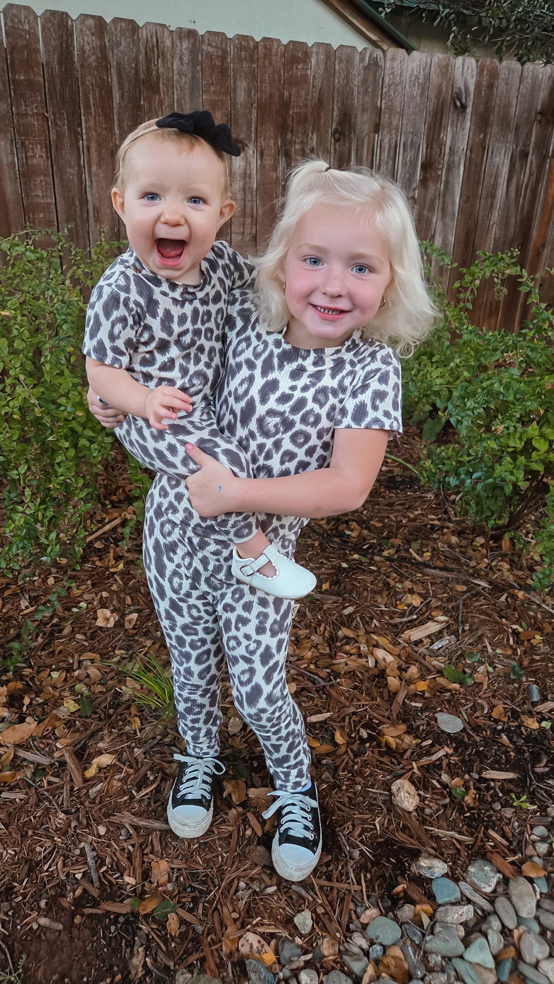 Two children in leopard print outfits standing outdoors with a wooden fence and greenery in the background.