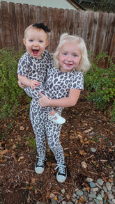 Two children in leopard print outfits standing outdoors with a wooden fence and greenery in the background.