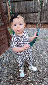 Baby in a leopard print outfit standing on a gravel surface with a wooden fence in the background.