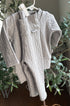 A light gray cable-knit baby romper with long sleeves and a mock neck, hanging on a white plastic hanger against a background of green plants.