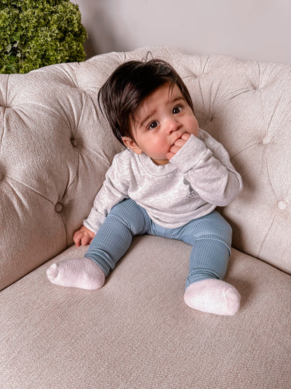 Baby sitting on a beige couch wearing a white shirt and blue pants.