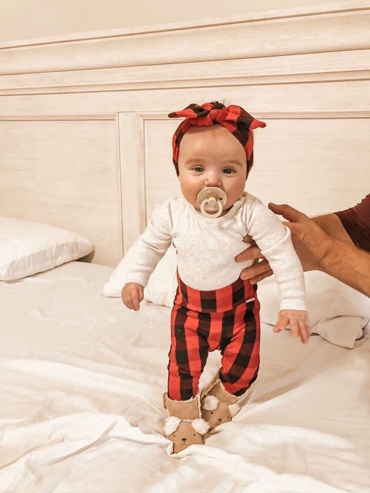 Sweet baby standing with support, dressed in a cozy red buffalo plaid headband and leggings, paired with a white onesie and fuzzy reindeer booties.