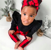 Adorable baby girl wearing a festive red buffalo plaid headband and leggings paired with a black ruffle sleeve top, sitting near mini frosted Christmas trees.