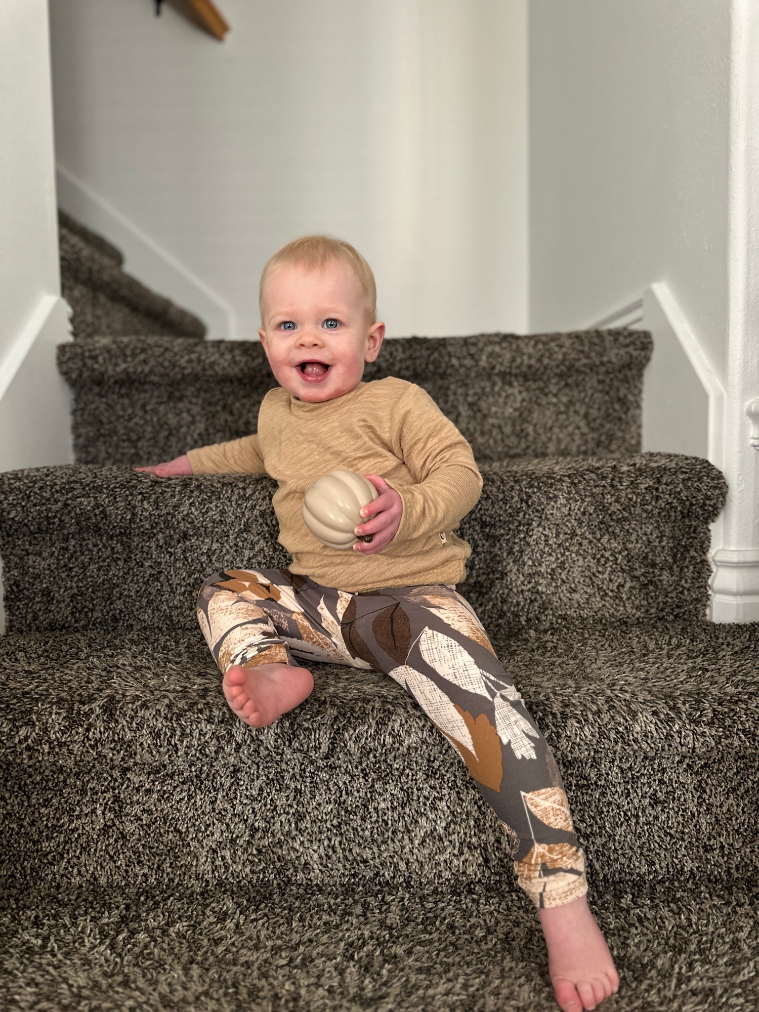 Baby sitting on a carpeted staircase holding a ball in Beige camel  textured sweater 