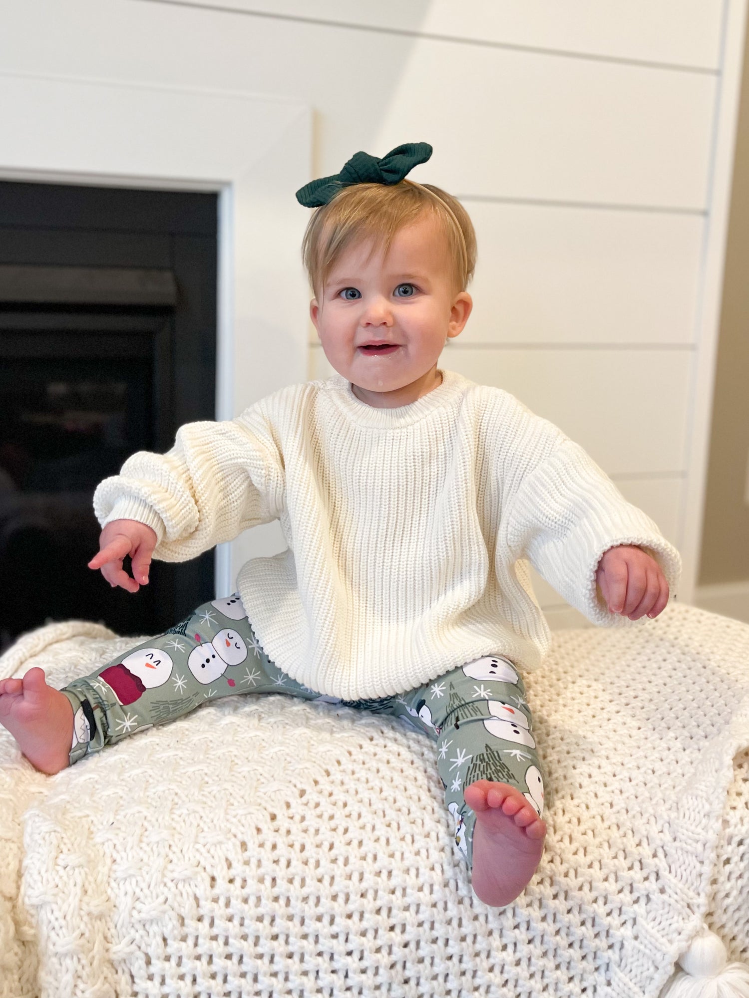 Baby sitting on a white surface wearing a white sweater and patterned pants with a green bow in hair.
