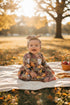 Baby wearing Handmade baby and toddler fall dress with scarecrow, pumpkins, corn, and autumn leaves pattern – Harvest Scarecrow print by Dudis Design. sitting on a blanket in a park with autumn leaves and a basket of apples.