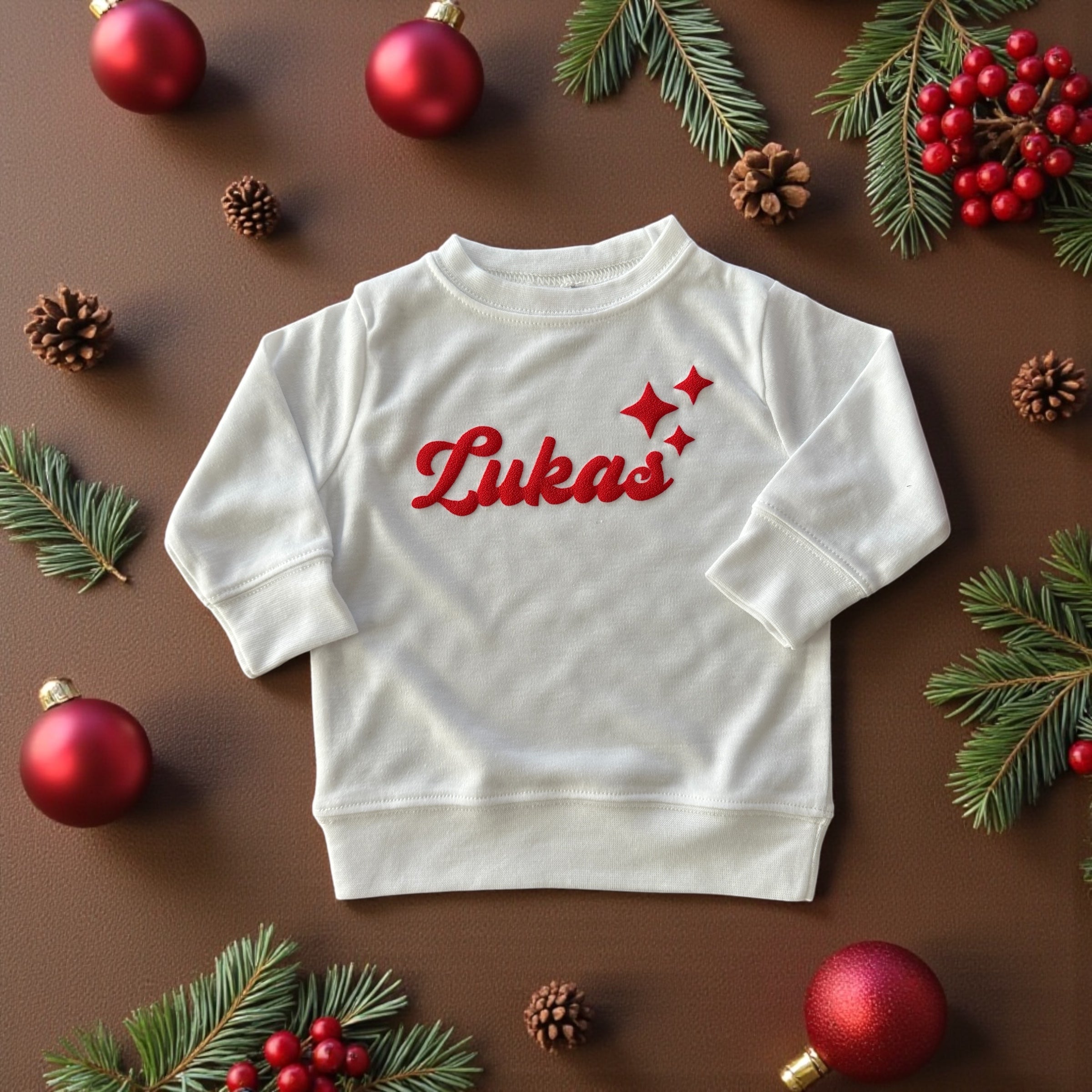 White baby sweatshirt with red “Lukas” name and star design, surrounded by Christmas ornaments and pine branches.