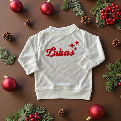 White baby sweatshirt with red “Lukas” name and star design, surrounded by Christmas ornaments and pine branches.