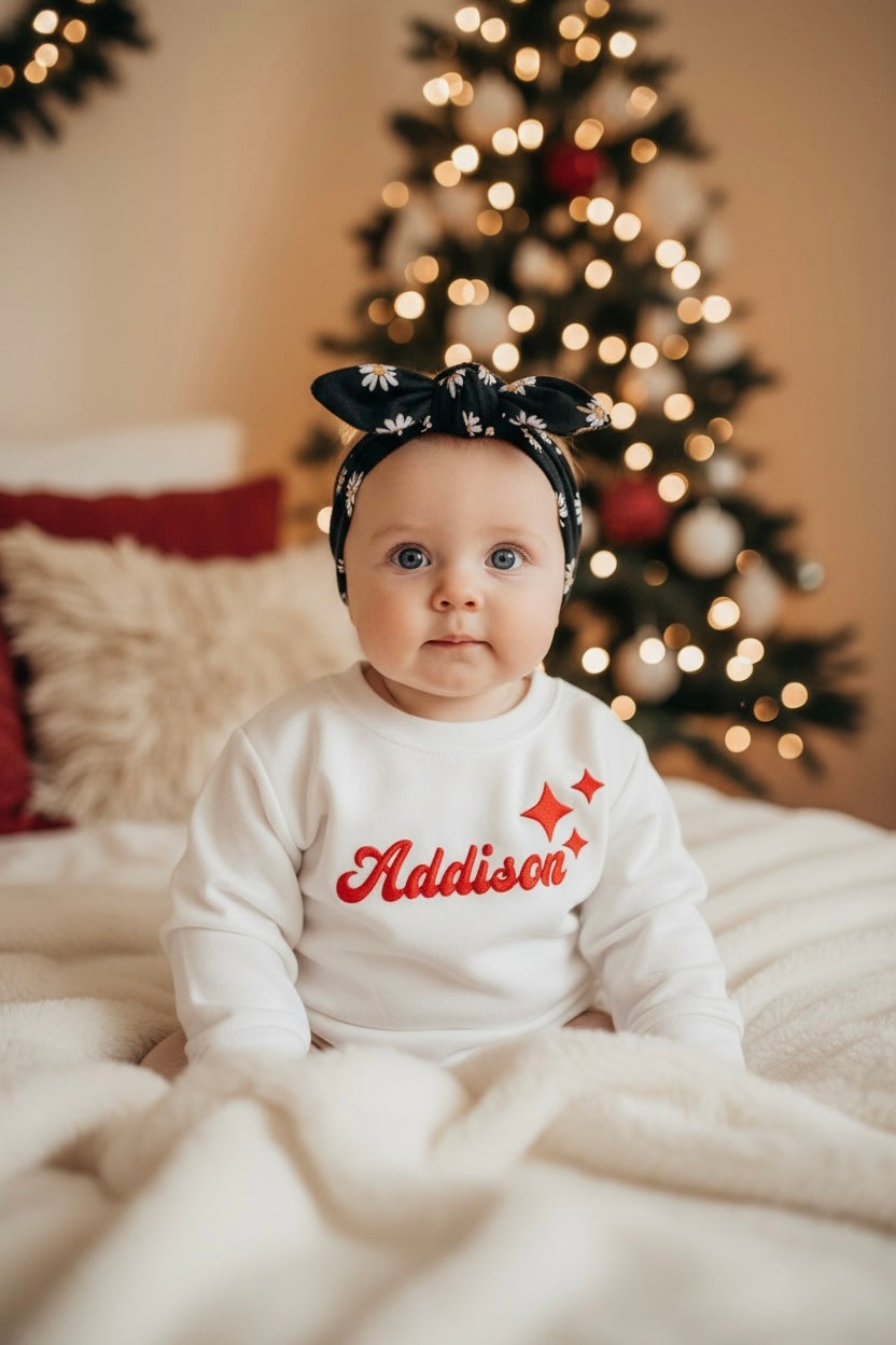 Baby wearing white personalized sweatshirt with red “Addison” name and star accents, sitting in front of a lit Christmas tree.