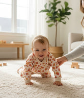 Smiling baby crawling on a cream carpet wearing a sunset leopard lounge set with coral and mustard print, supported gently by a parent in a bright cozy nursery.