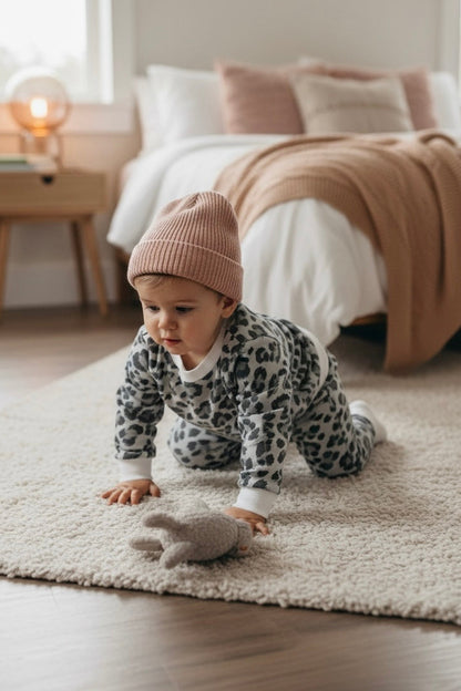 Baby in leopard print outfit and pink beanie playing with a toy on a rug in a bedroom.