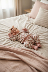Newborn baby peacefully sleeping on a bed, wearing a dusty pink checkered lounge set with matching cuffs and a soft bow headband; warm neutral bedroom tones in the background.