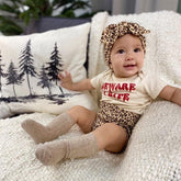 Baby wearing Leopard Print Bummies and matching leopard headband, paired with a "Beware of Cute" graphic onesie and beige knee-high socks, seated on a cozy knit blanket with a woodland-themed cushion in the background.