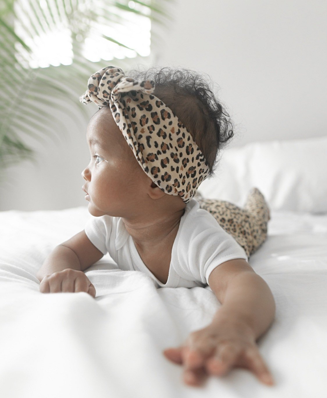 Baby wearing a Tan Glitter Cheetah top knot headband, lying on a white surface with natural lighting and greenery in the background, showcasing a trendy handmade baby accessory.