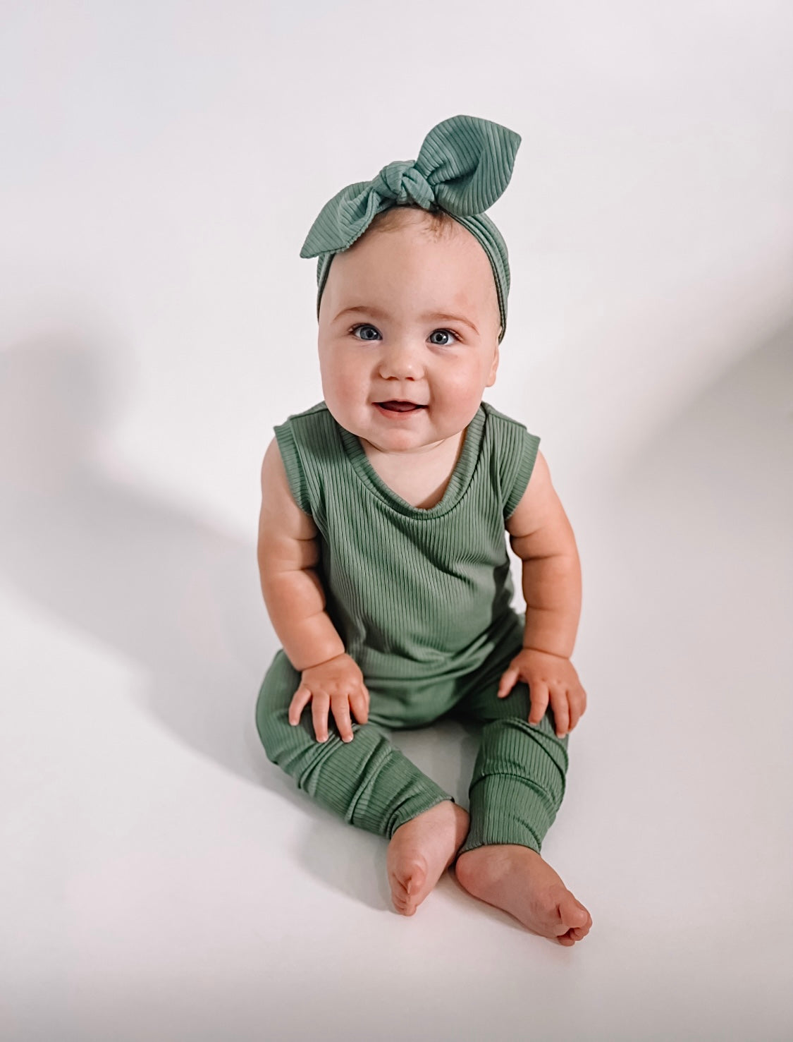 A smiling baby wearing a sleeveless ribbed green romper paired with a matching bow headband, sitting on a neutral background.