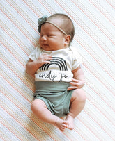 Adorable newborn baby wearing ribbed green bummies, a custom rainbow onesie, and a matching bow headband, peacefully sleeping on a striped blanket.