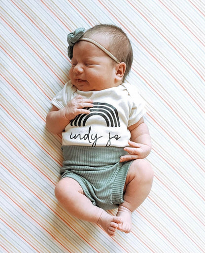 Adorable newborn baby wearing ribbed green bummies, a custom rainbow onesie, and a matching bow headband, peacefully sleeping on a striped blanket.