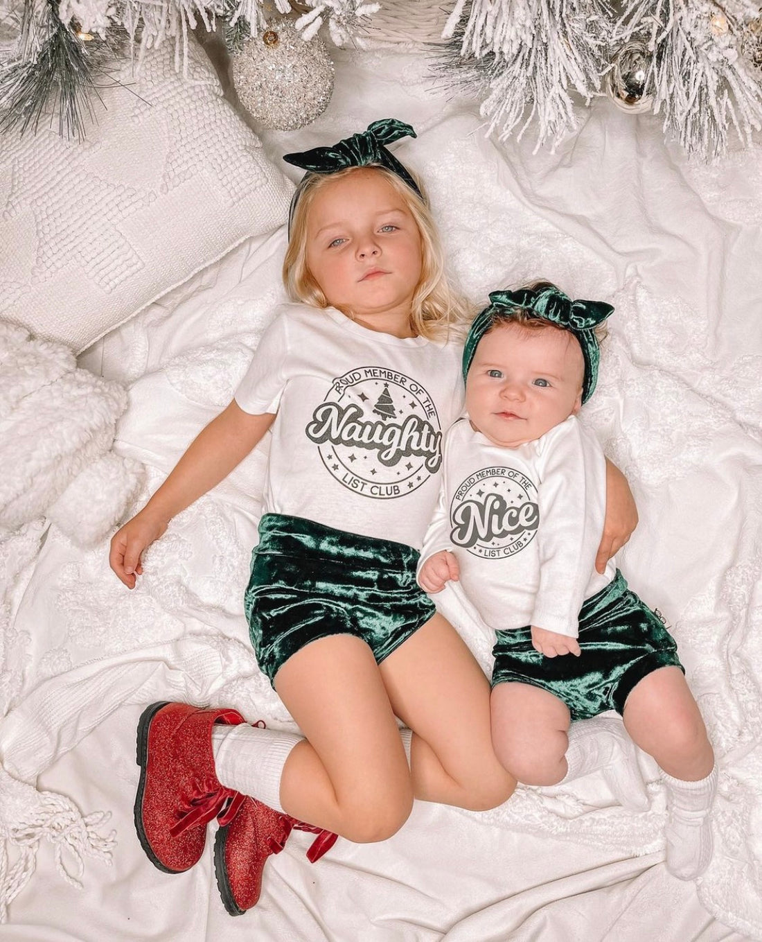 Siblings lying under a decorated Christmas tree, wearing emerald green velvet bummies and matching headbands with &quot;Naughty&quot; and &quot;Nice&quot; themed holiday shirts. A charming coordinated outfit set.