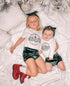 Siblings lying under a decorated Christmas tree, wearing emerald green velvet bummies and matching headbands with "Naughty" and "Nice" themed holiday shirts. A charming coordinated outfit set.